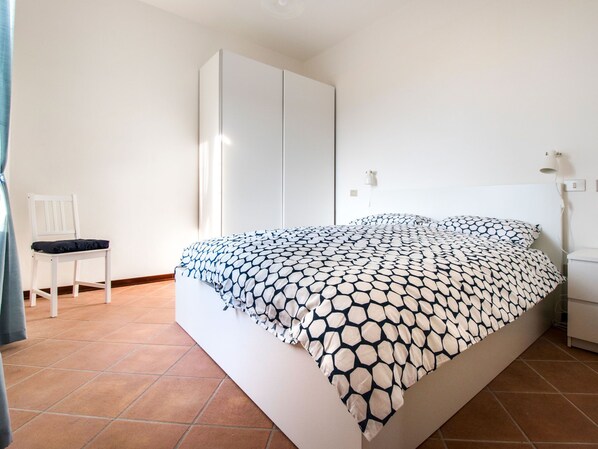 A bedroom with a bed, a white wardrobe, a chair, and a bedside table.