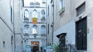 A narrow street with a building featuring a balcony with potted plants, a sign with a shield emblem, and a black door with a number plaque. - Morgana (FLG208) by Interhome (Finale Ligure)
