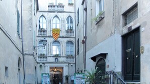 Una calle estrecha con un edificio que tiene un balcón con plantas en macetas, un letrero con un escudo y una puerta negra con placa numérica.