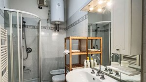 Badeværelse med glasvæg omkring bruseniche, toilet, vask med spejl og en træhylde med toiletartikler.