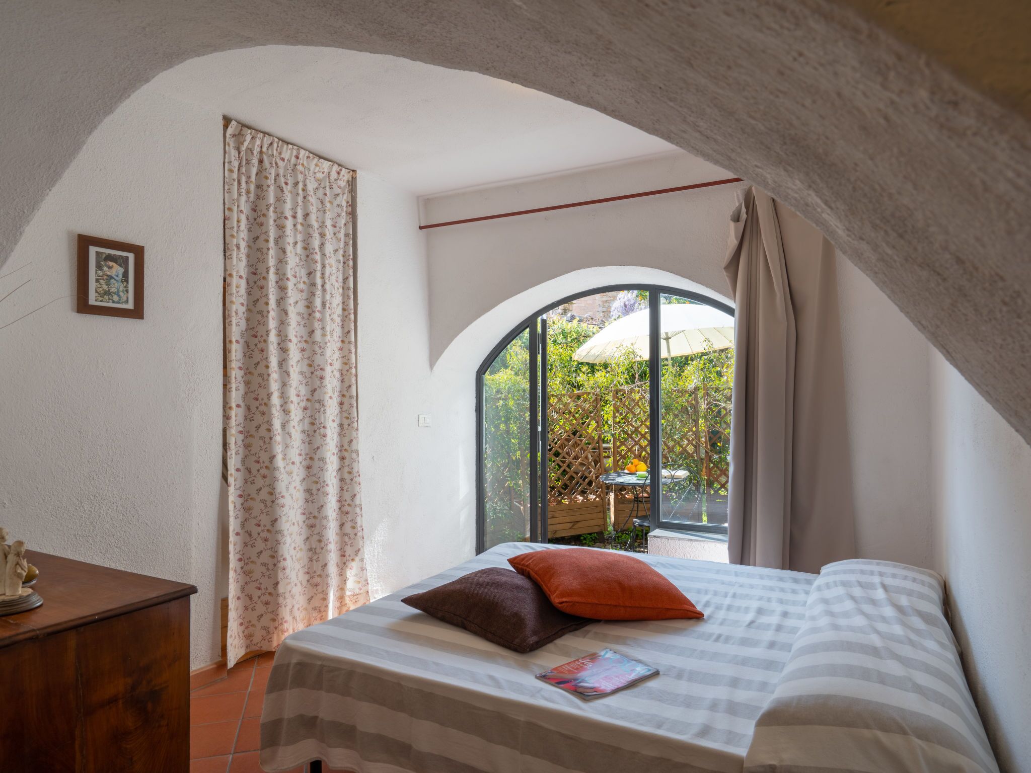 Ein Schlafzimmer mit einem Bett, einer Holzkommode, einem gerahmten Bild und einem Fenster mit Blick auf Grün.