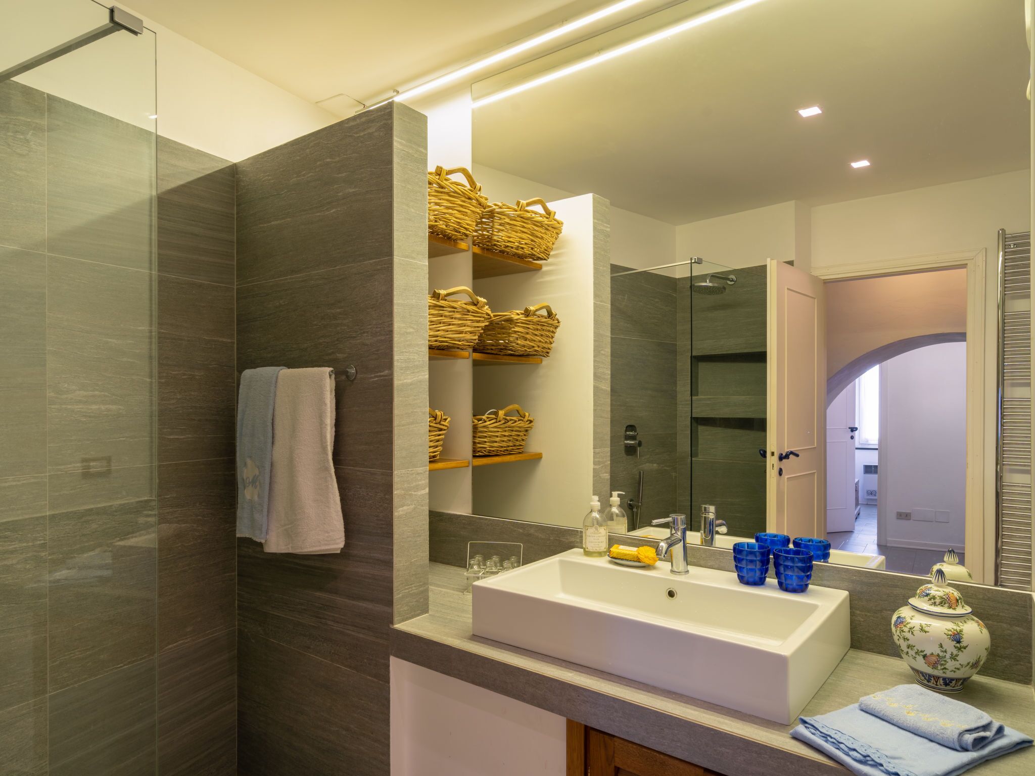 A modern bathroom with a glass shower enclosure, a white sink, and wicker baskets on shelves.