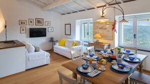 Una stanza ben illuminata con soffitto in legno, pareti bianche, un divano, un tavolo da pranzo apparecchiato con piatti bianchi e azzurri e quadri appesi al muro.