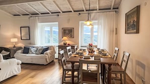 A cozy room with a wooden dining table set for four, a sofa, a lamp, and a view of the outdoors through large windows. - Laura (DOL281) by Interhome (Dolcedo)
