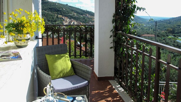 A balcony with a wicker chair, a table with tea set, and a view of a hilly landscape. - Villa Paradiso (DOL192) by Interhome (Dolcedo)