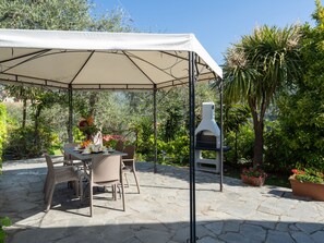 Eine Terrasse im Freien mit Tisch und Stühlen, einem Pizzaofen und Kübelpflanzen, alles unter einem Sonnensegel.