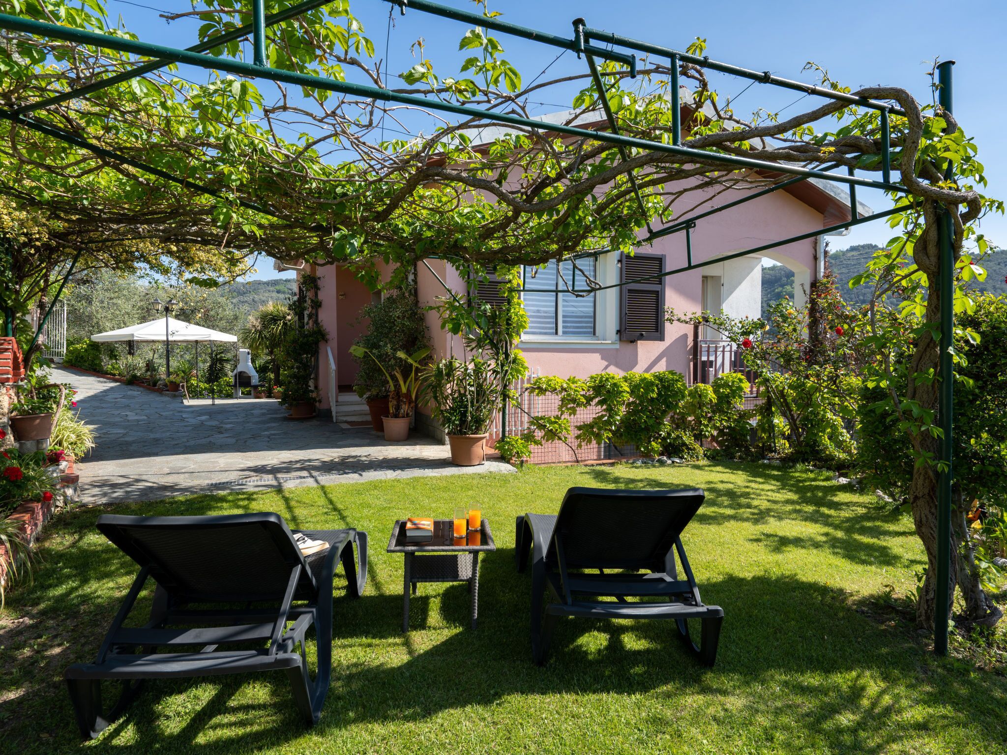 A patio with two black lounge chairs and a table, surrounded by potted plants and a building with a pink facade.