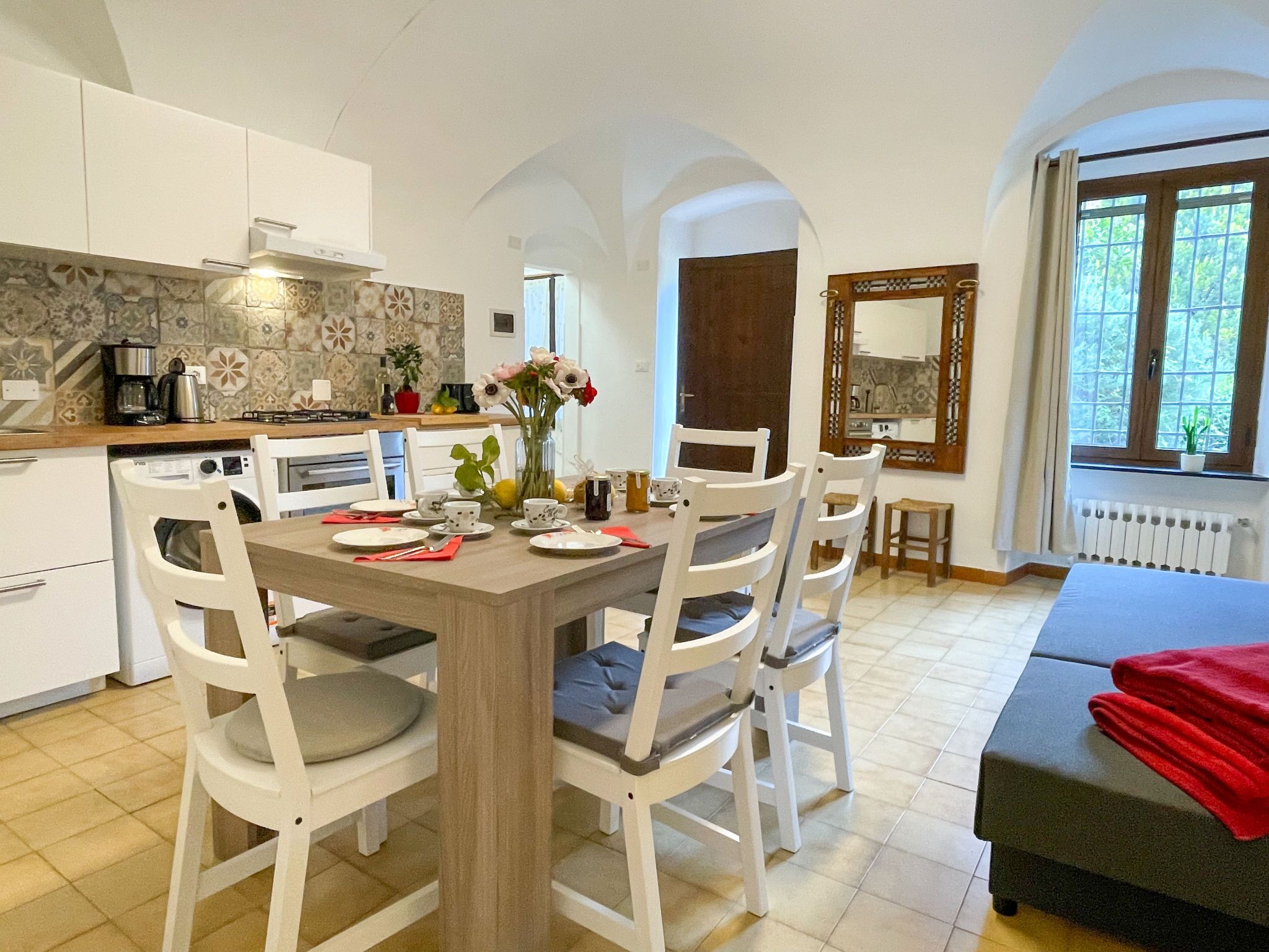 A dining area with a wooden table and white chairs, a kitchen with a tiled backsplash, and a window with a view of greenery.