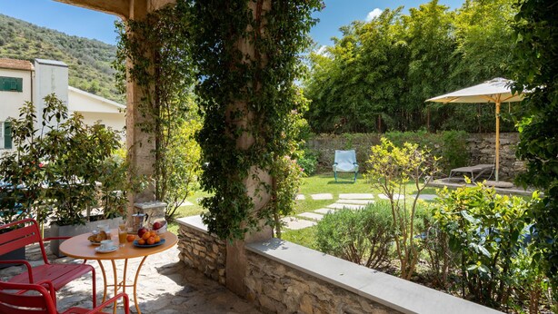 Un espace extérieur aménagé avec un mur en pierre, une table entourée de chaises et une vue sur un jardin et des collines en arrière-plan.