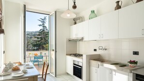 Une cuisine moderne avec des meubles blancs, une cuisinière à gaz et un évier. On y trouve un coin repas avec une table et des chaises, et une vue sur les arbres à travers une porte vitrée.