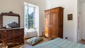 Une chambre avec une commode en bois, un grand miroir, une fenêtre donnant sur des immeubles, une armoire en bois, un lit recouvert d’une couvre-lit à motifs et une chaise.