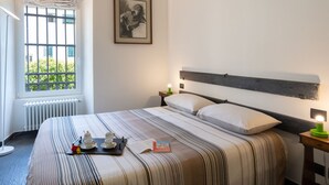 Une chambre d’hôtel avec un lit, une fenêtre grillagée, un tableau encadré au mur et une table de chevet avec une lampe.