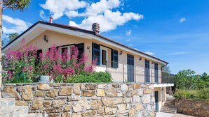 A two-story house with a stone wall in front, a balcony with black railings, and pink flowering plants. - Settimo Cielo (DOL400) by Interhome (Dolcedo)