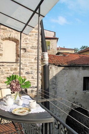 A balcony with a table set for tea, a vase with flowers, and a view of a stone building with a tiled roof.