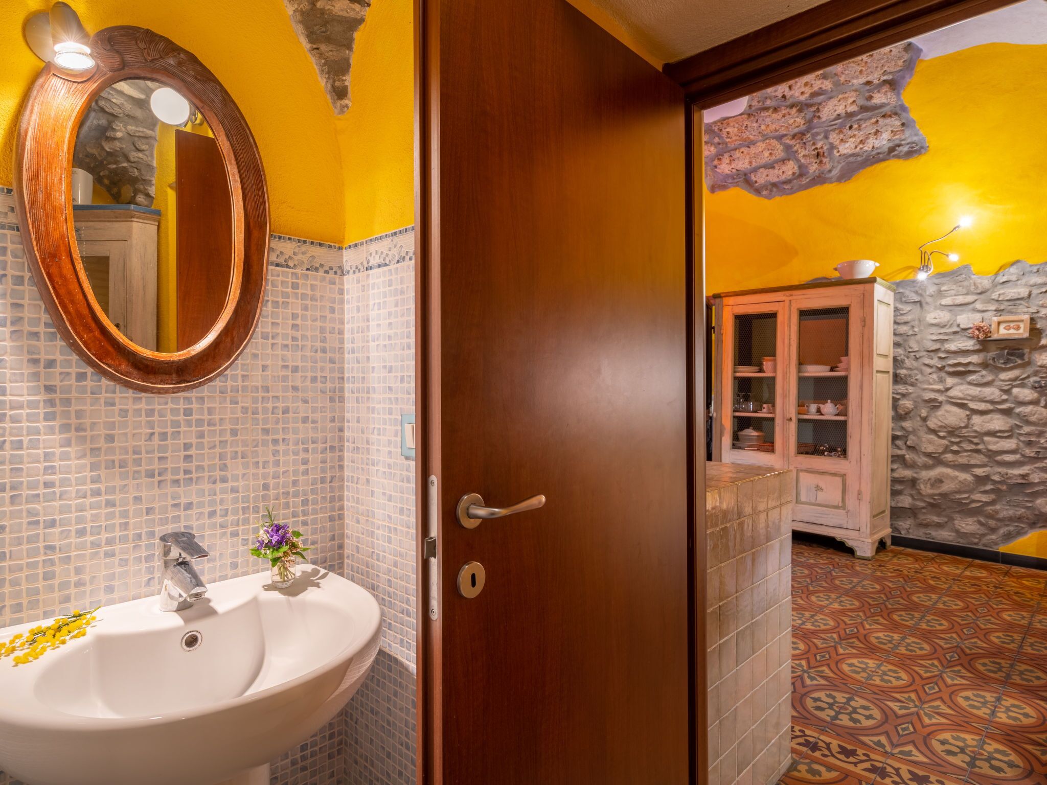 A bathroom with a white sink, a mirror, and a door leading to a room with a stone wall and a cabinet.