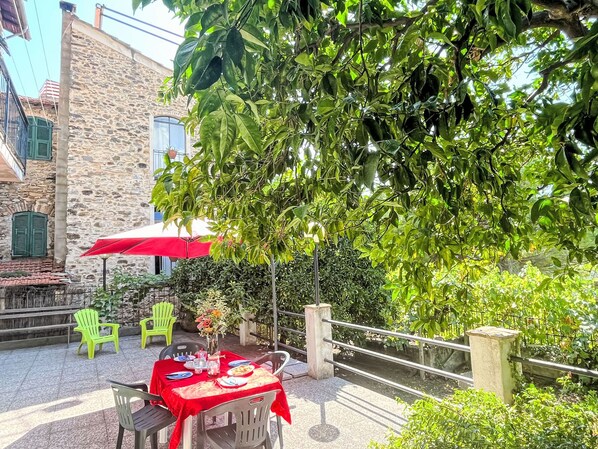 Un espace de restauration en plein air avec une table dressée pour deux personnes, placée sous un parasol rouge, entourée de verdure et d’un bâtiment en pierre.