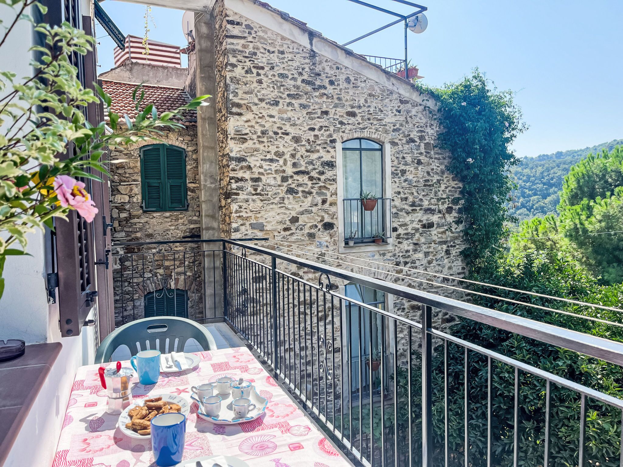 Ein Balkon mit einem Tisch, gedeckt für Tee, mit Blick auf ein Steinhaus und üppiges Grün.