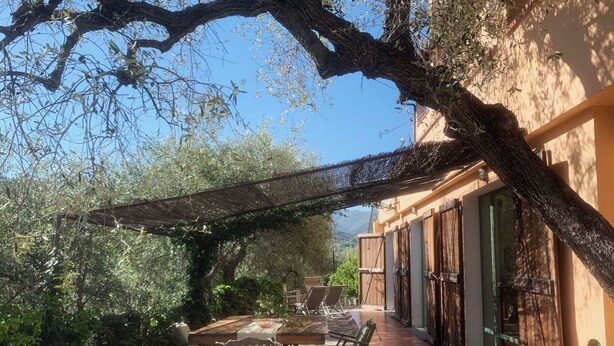 An outdoor patio with a table and chairs, surrounded by greenery and a building with large windows. - Apartment NISSO by Interhome (Dolcedo)