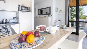 A modern kitchen with white cabinets, a microwave, and a refrigerator. A dining table with a colorful plate of fruits and a white mug is in the foreground. - Le Margherita (SLR266) by Interhome (San Lorenzo al Mare)
