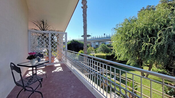 A balcony with a table and chairs, a white railing, and a view of a bridge and greenery. - Villa Gisi (Fiumicino)