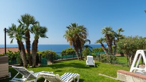 Área externa gramada com cadeiras de lounge brancas e vista para o mar.