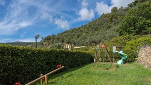 Ein grünes Freigelände mit Schaukel und Rutsche, umgeben von einer Hecke und einer Steinmauer.