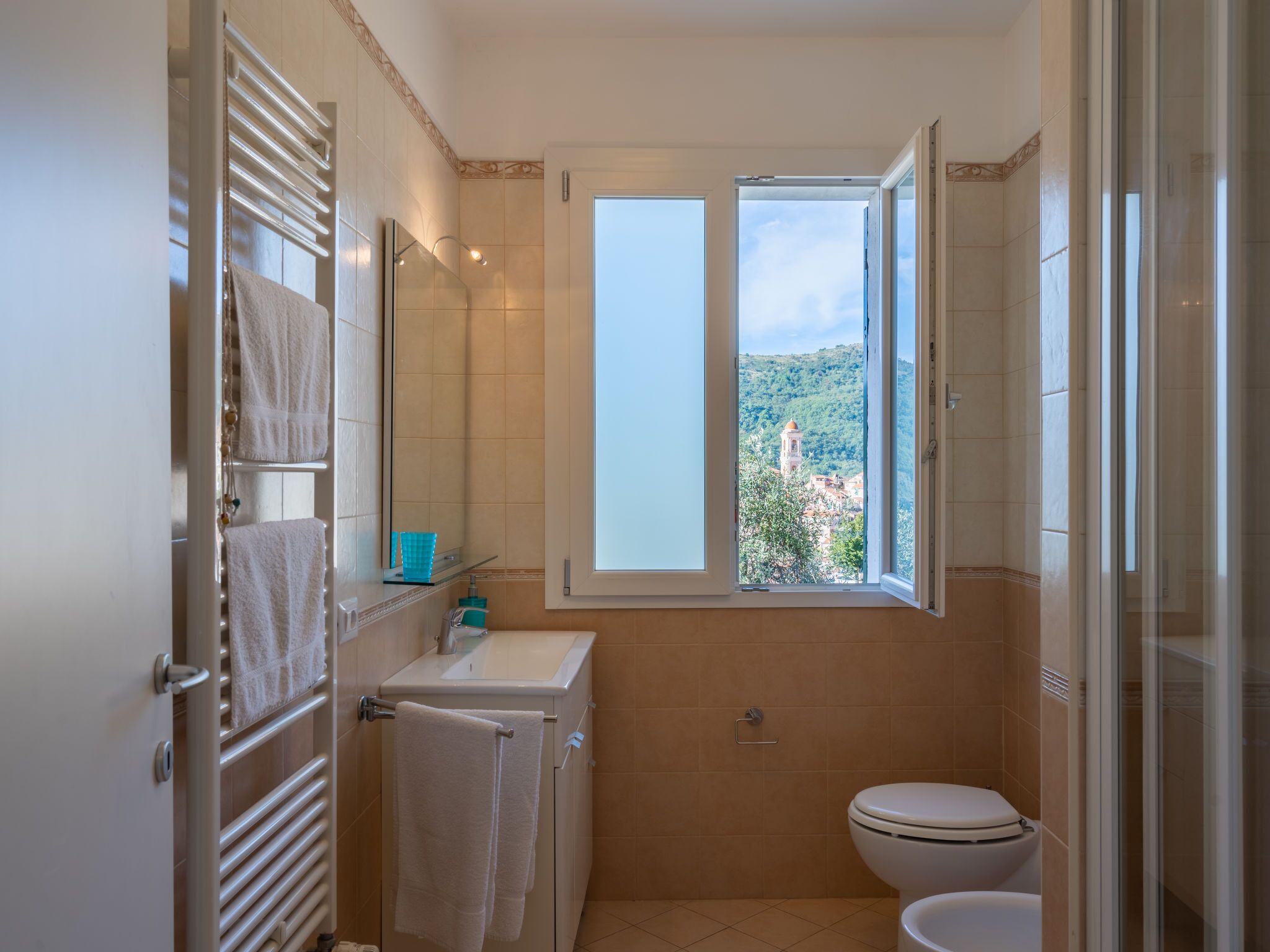 A bathroom with a toilet, sink, and towel rack. There is a window with a view of a hill and buildings.