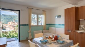 Un comedor con una mesa preparada para el desayuno, vistas de un pueblo a través de una ventana y una cocina con armarios de madera.