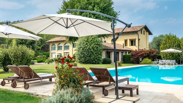 A poolside area with lounge chairs and umbrellas, a well-maintained lawn, and a large house in the background. - Villa Carlotta by Interhome (Costigliole d'Asti)