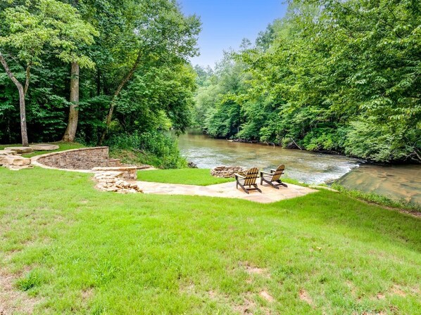 Een terras aan de rivier met Adirondack-stoelen, een stenen muur en weelderige begroeiing.