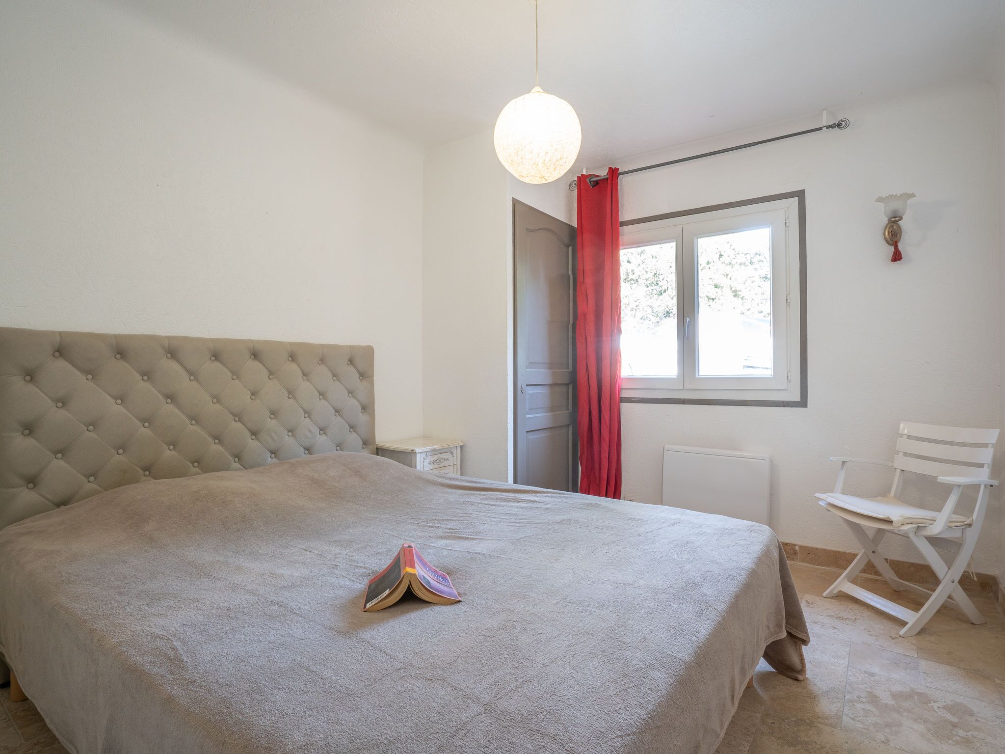 Une chambre à coucher avec une tête de lit capitonnée, un lit recouvert d’une couverture beige, une petite table de chevet, une fenêtre avec un rideau rouge et une chaise blanche.