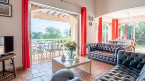 A spacious living room with a leather sofa, a coffee table, and a TV. There is a balcony with red curtains and a dining area with a table and chairs. - Bartavelle by Interhome (Sainte Maxime)