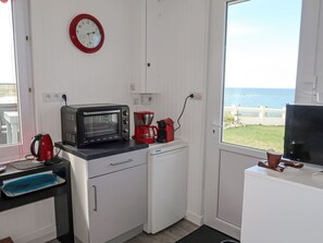 A compact kitchen with a microwave, coffee maker, kettle, and a small refrigerator. A clock is mounted on the wall, and a window offers a view of the sea.
