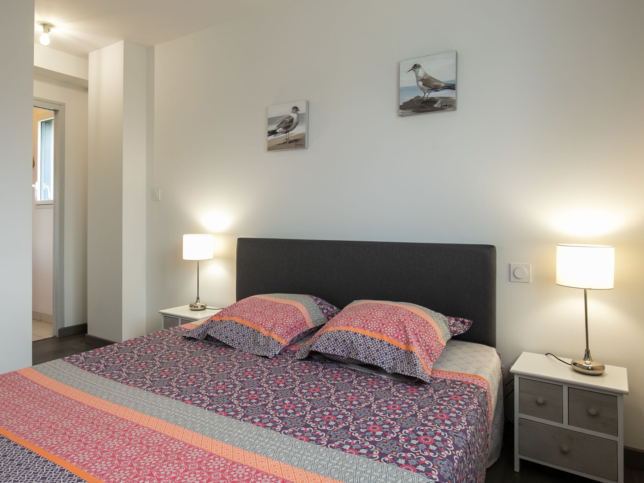 A neatly made bed with patterned bedding, two wall-mounted lamps, and two framed artworks on the wall.
