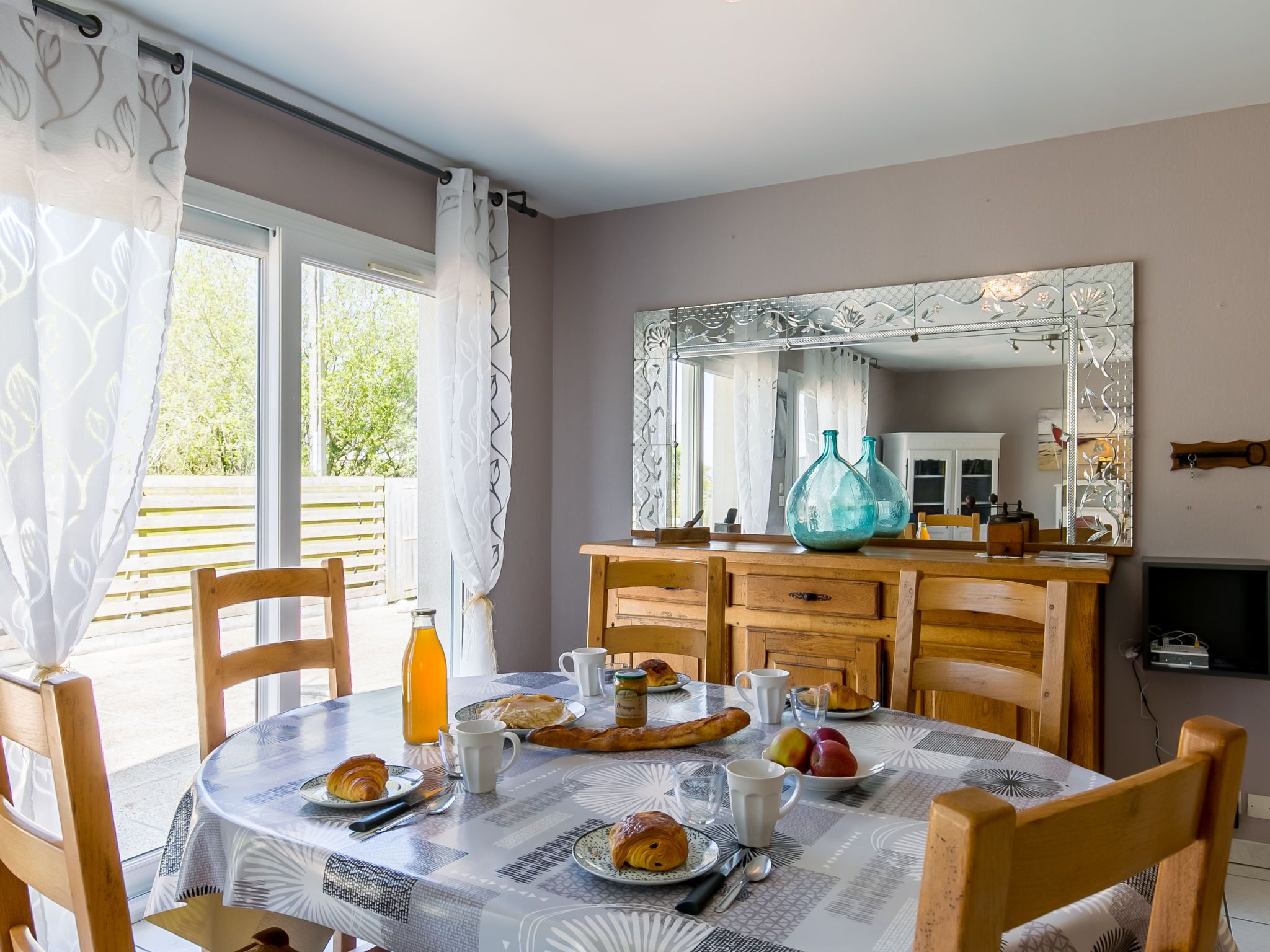 A dining area with a round table set for breakfast, featuring croissants, fruits, and beverages.