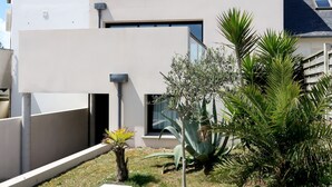 Un edificio moderno de dos pisos con techo plano, ventanas amplias y un pequeño jardín con diversas plantas.