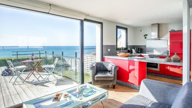 Un apartamento moderno con balcón que da al mar, una cocina con armarios rojos y una sala de estar con una mesa de centro de vidrio.