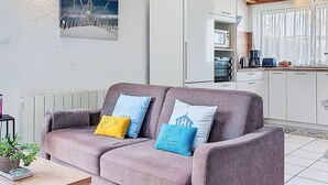 A cozy living room with a sofa, a painting, and a kitchen area with cabinets and appliances. - Tal Ar Mor by Interhome (Plouescat)