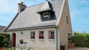 A two-story house with a sloped roof, a chimney, and a small dormer window. There are flower boxes on the windowsills and a small table with chairs in the front yard. - Tal Ar Mor by Interhome (Plouescat)