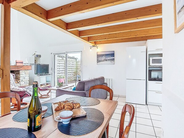 A cozy kitchen with wooden beams, a dining table with croissants, a bottle of beer, and a TV in the background.