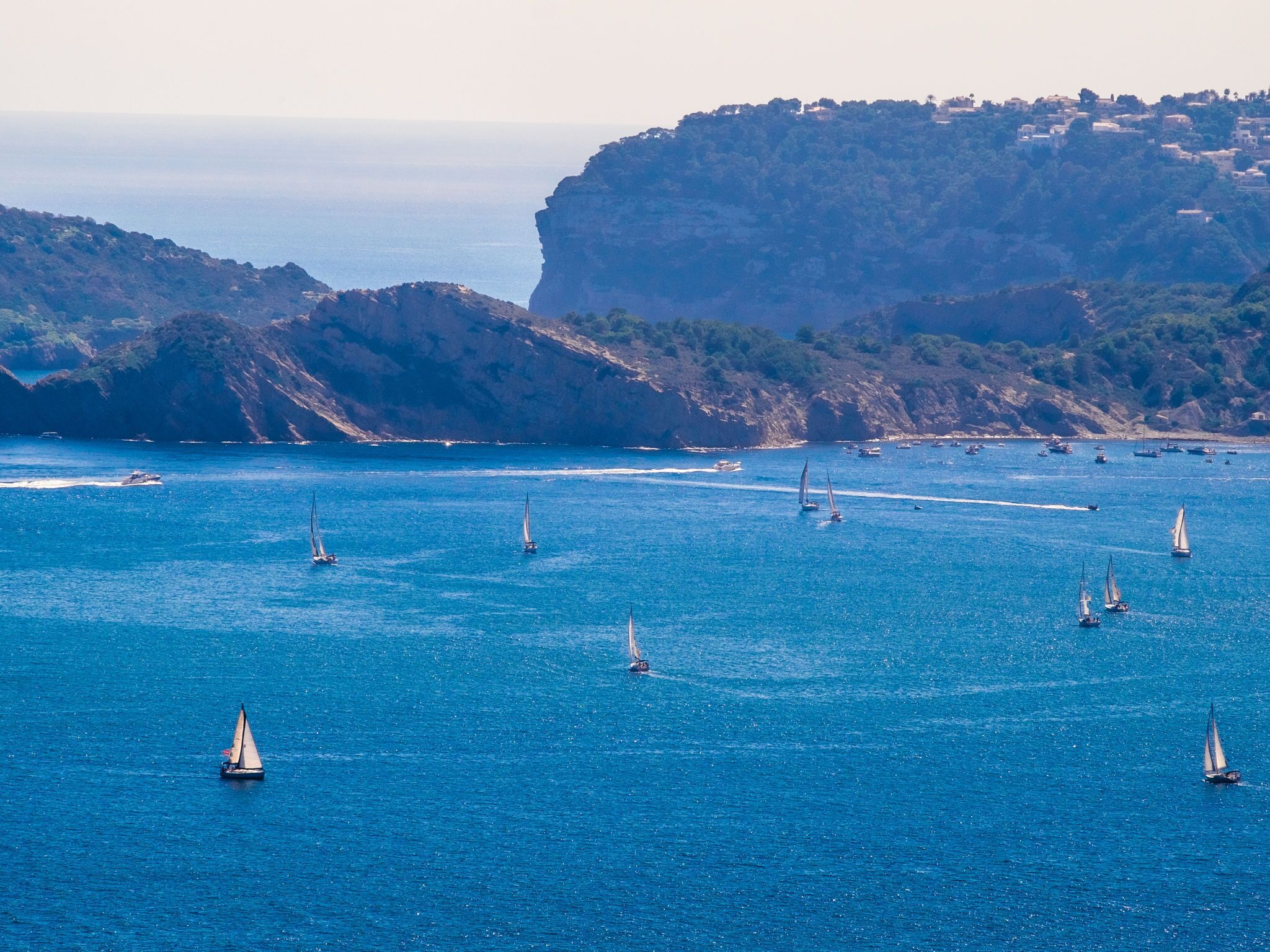 A coastal scene with multiple sailboats on the water, surrounded by rocky cliffs and a distant town.