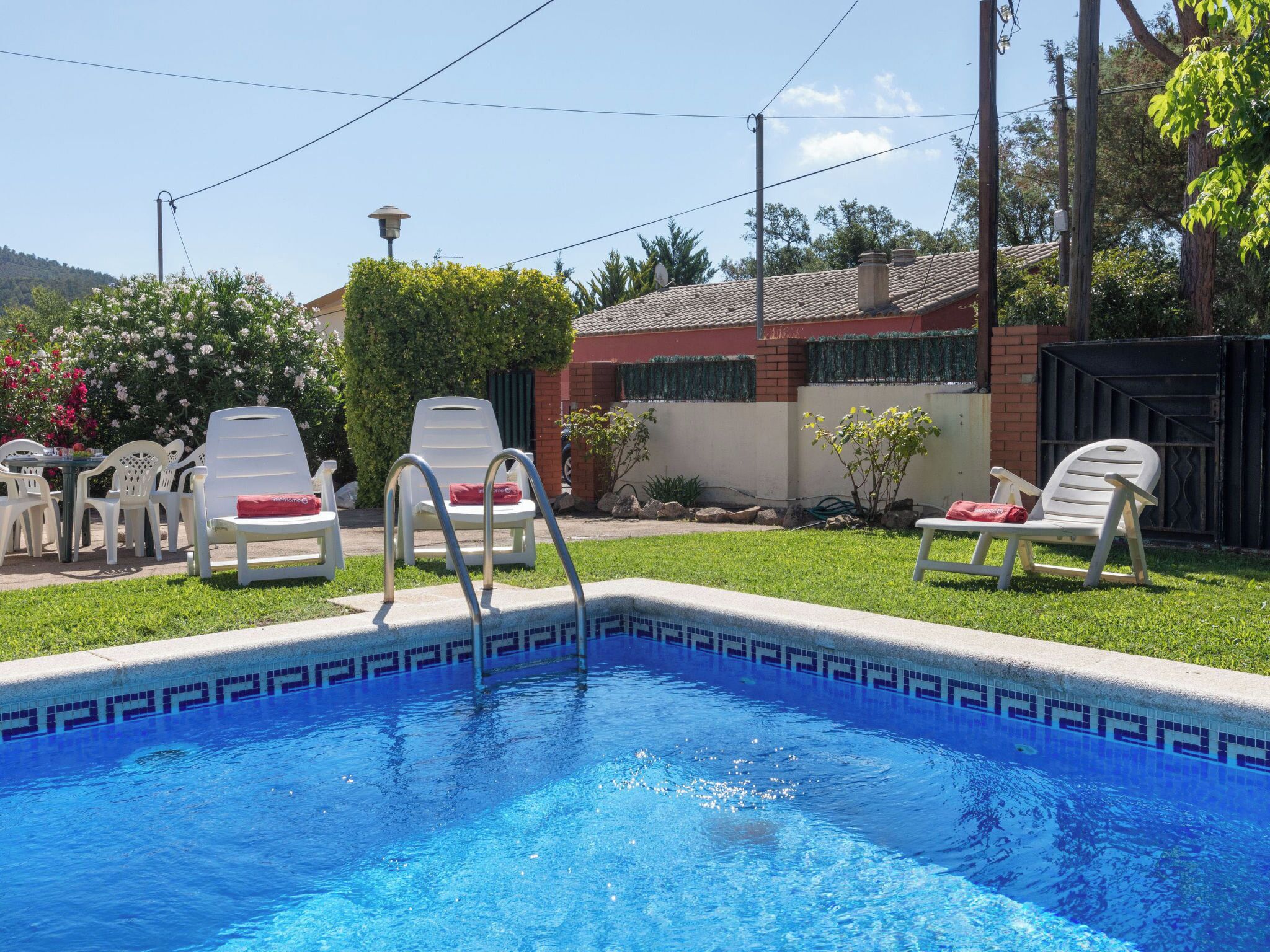 Una piscina con sillas de descanso y una pequeña mesa, rodeada de césped y un edificio con tejado de baldosas.