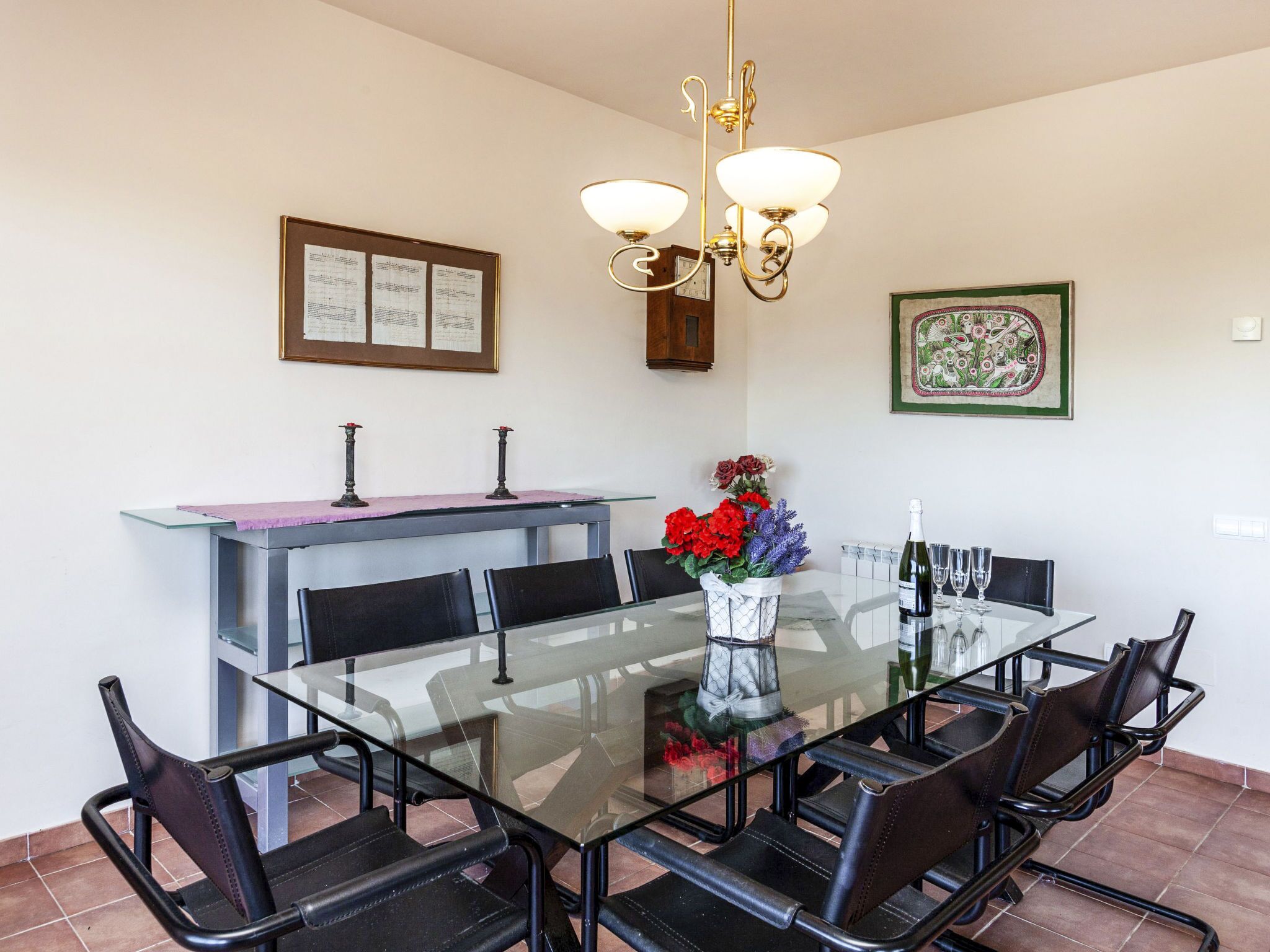 A dining area with a glass table, black chairs, a vase with flowers, a bottle of wine, and a chandelier.