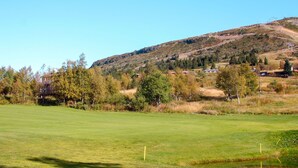 A golf course with a clear view of the surrounding landscape, including a hill with sparse vegetation and a few buildings in the distance.