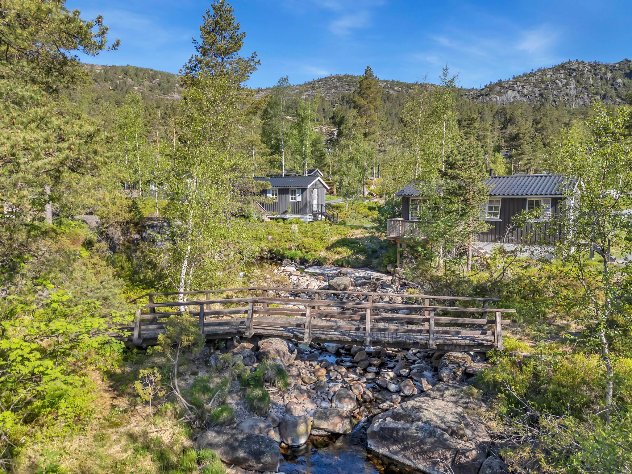 A rustic cabin with a wooden deck and a small bridge over a stream, surrounded by dense forest and hills.