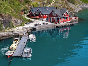 A red house with a dock extending into a calm body of water, boats moored at the dock, and a path leading to the house.