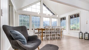 Amplia habitación con techo alto, ventanas grandes que ofrecen vistas a la montaña, zona de comedor con mobiliario de madera y un sillón tapizado en gris con un reposapiés a juego.