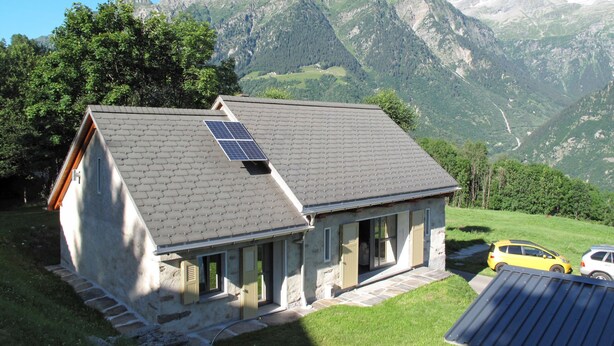 Un bâtiment en pierre doté d’un toit en pente et d’un panneau solaire, entouré d’une végétation luxuriante et de montagnes.