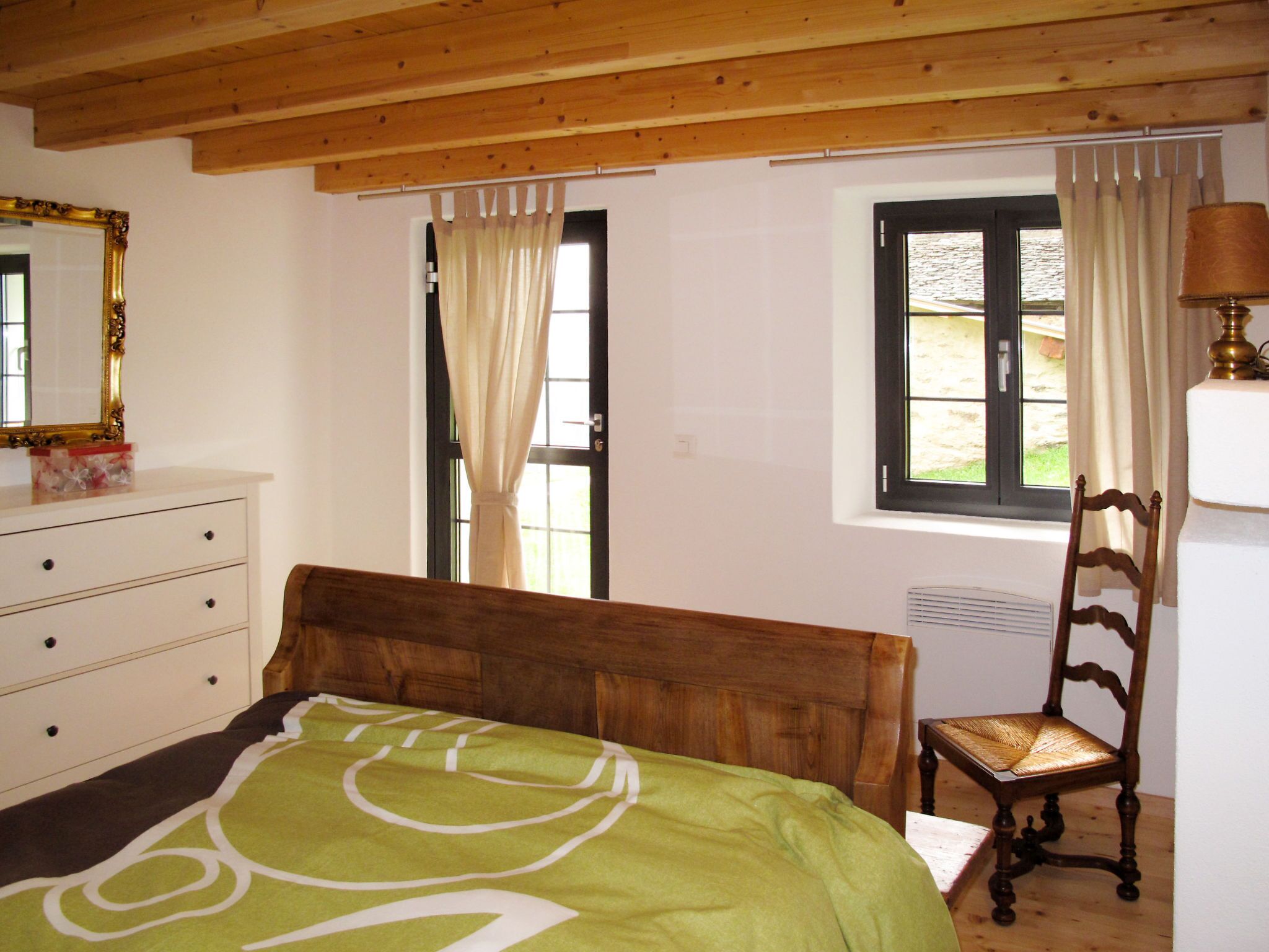 A bedroom with a wooden headboard, a wooden chair, a bed with a green blanket, a dresser, a mirror, and a window with curtains.
