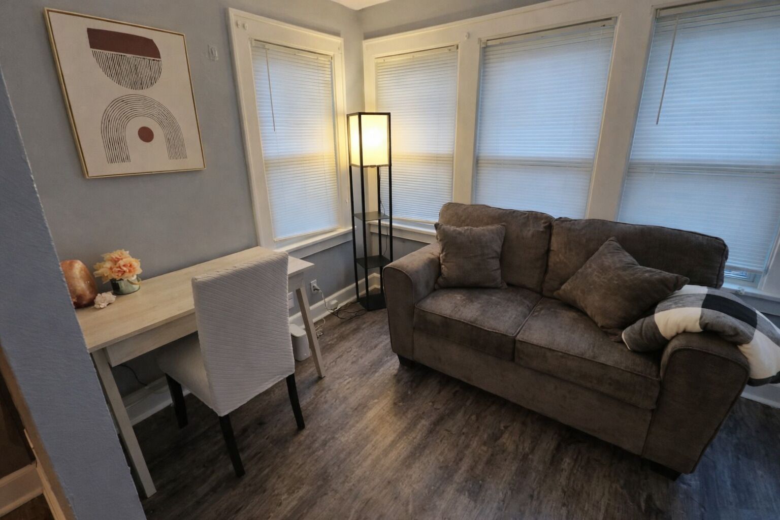 A living room with a brown sofa, a white chair, a desk, and a floor lamp.